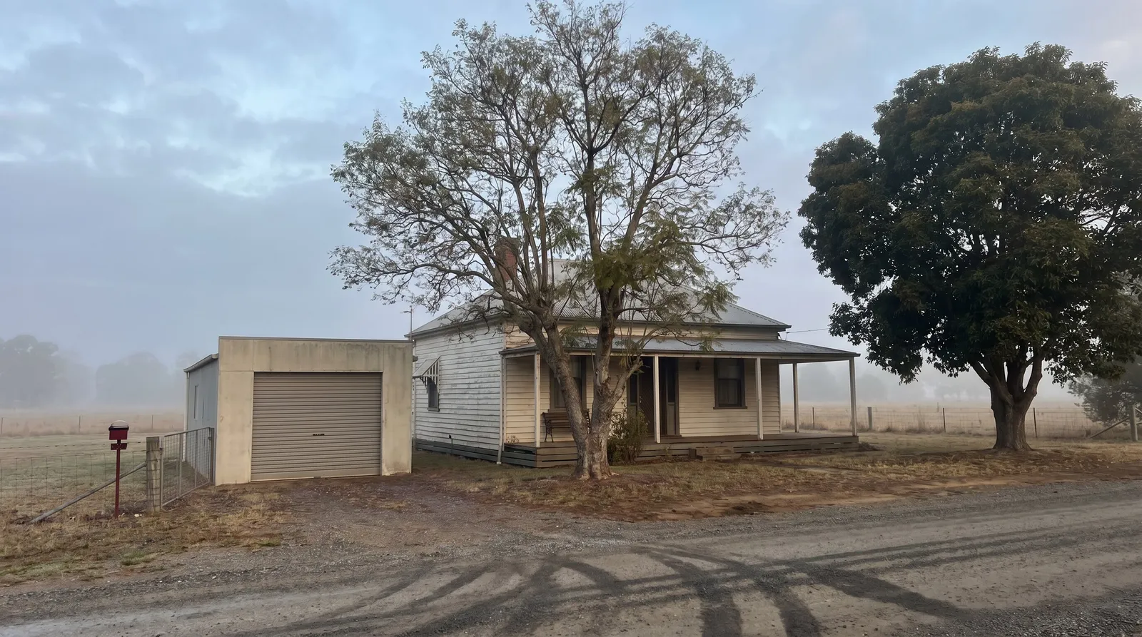 An older weatherboard cottage and its detached garage with a dated roller door, morning fog over the river flats behind